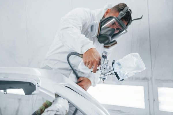 In protective mask. Painting the car. Caucasian automobile repairman in uniform works in garage In protective mask. Painting the car. Caucasian automobile repairman in uniform works in garage.
